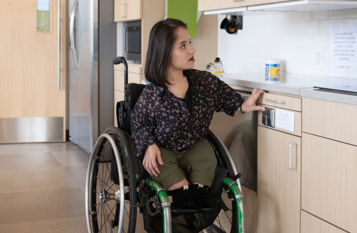 A South Asian person sits in her wheelchair in an accessible kitchen - Disabled And Here.